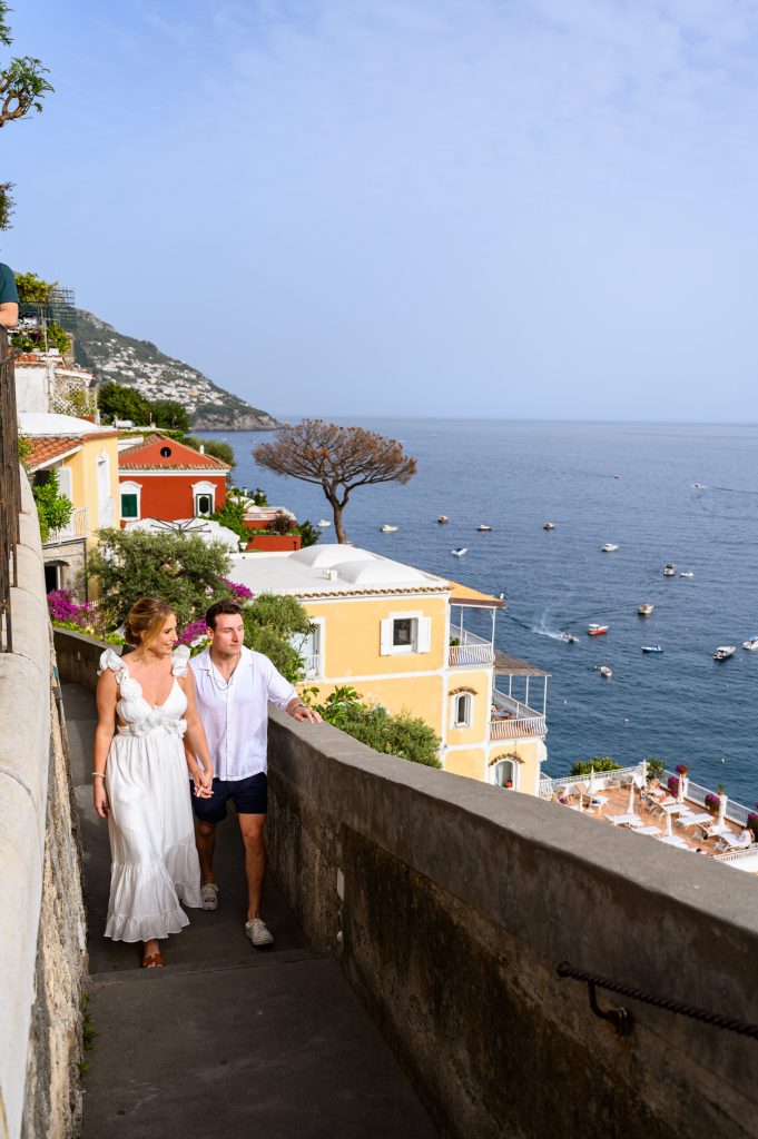 Couple walking hand in hand through Positano with colorful buildings and sea view in the background during engagement session
