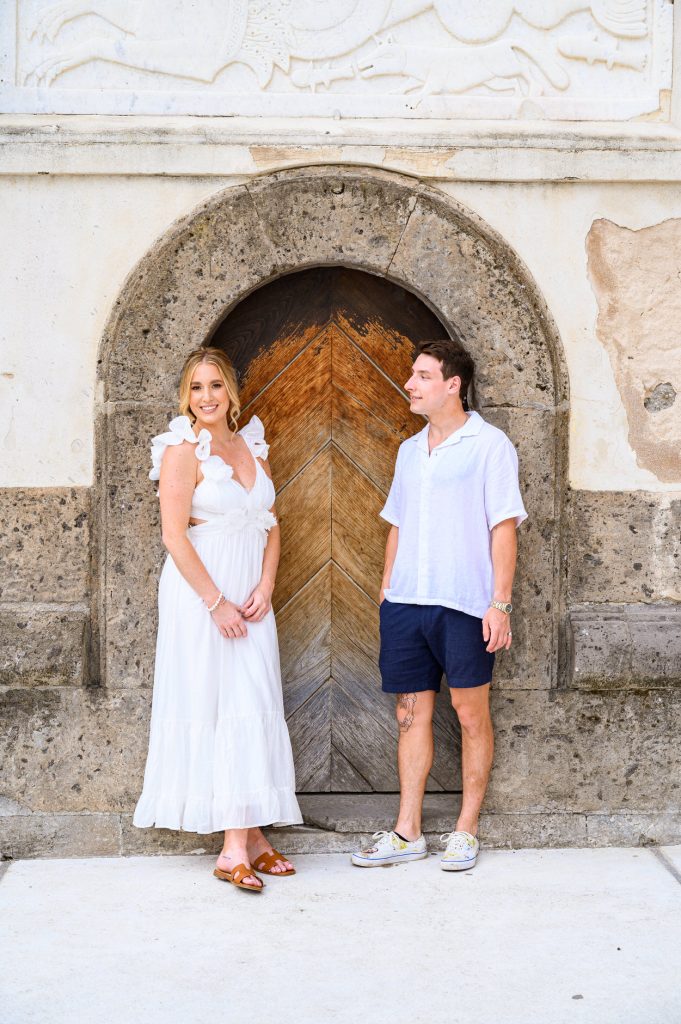 Engaged couple smiling and standing in front of an ancient arched stone doorway with carved details in Positano.