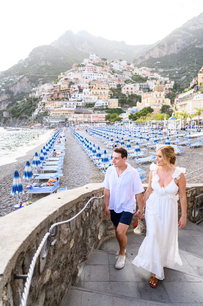 Engaged couple walking hand in hand along the beach promenade in Positano with colorful buildings and blue beach umbrellas in the background.