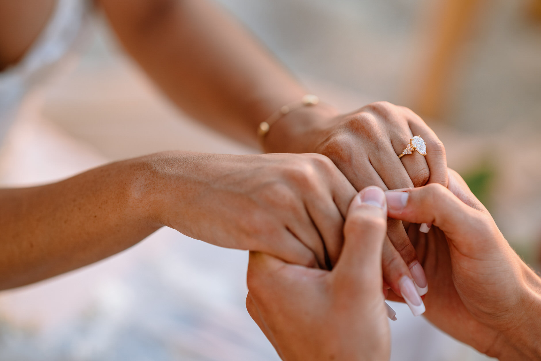 An intimate close-up shot of the newly engaged couple's hands clasped together, featuring the engagement ring, on a sunset beach with a scenic view.