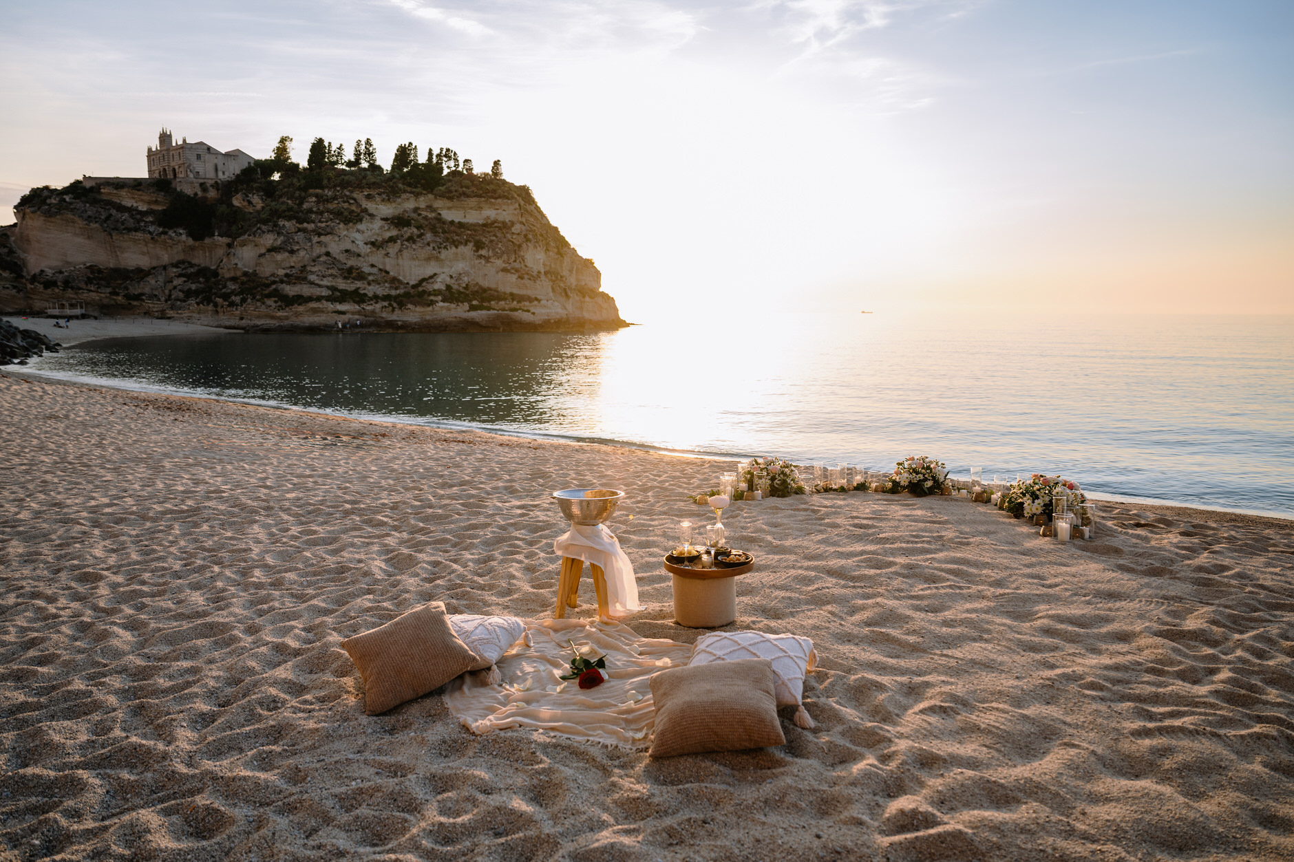 Romantic beach picnic setup in Tropea featuring woven cushions, flowers, and champagne